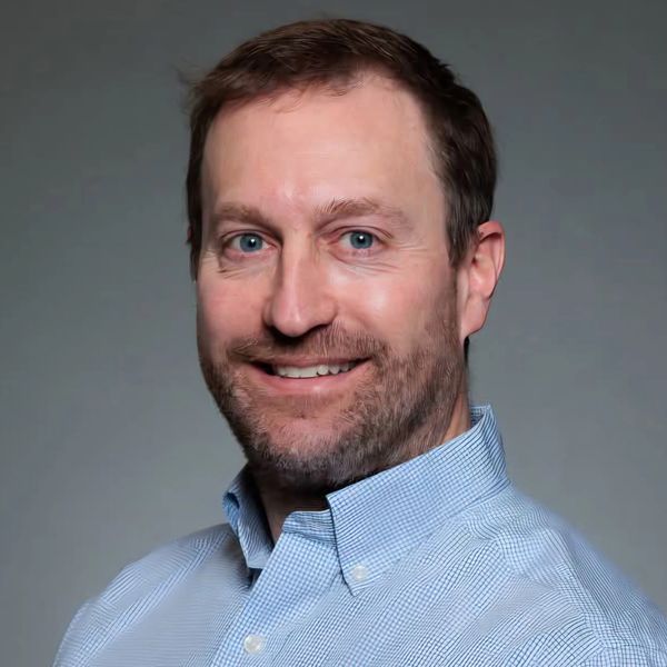 Smiling man in a light blue checkered shirt against a gray background.