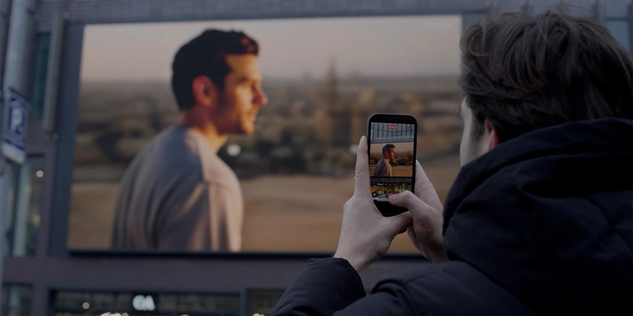 Person taking photo of a large digital billboard showing a man in profile.