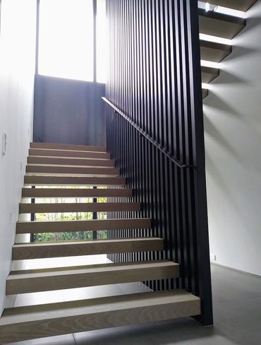 Open timber treads rise beside tall black metal balusters and a slim handrail in a bright interior.