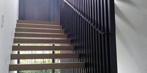 Open timber treads rise beside tall black metal balusters and a slim handrail in a bright interior.