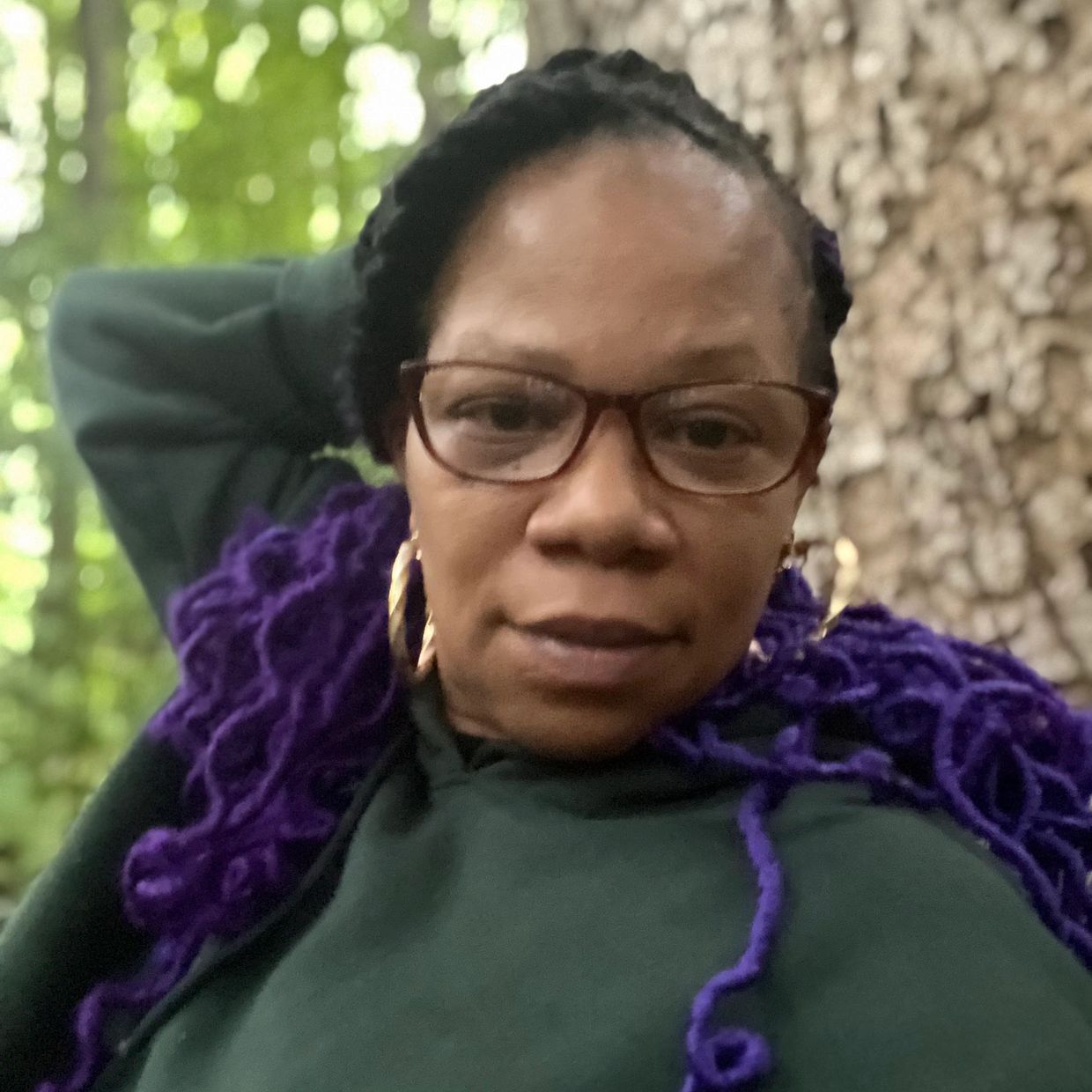 Woman with purple braided hair and glasses poses outdoors near a tree.