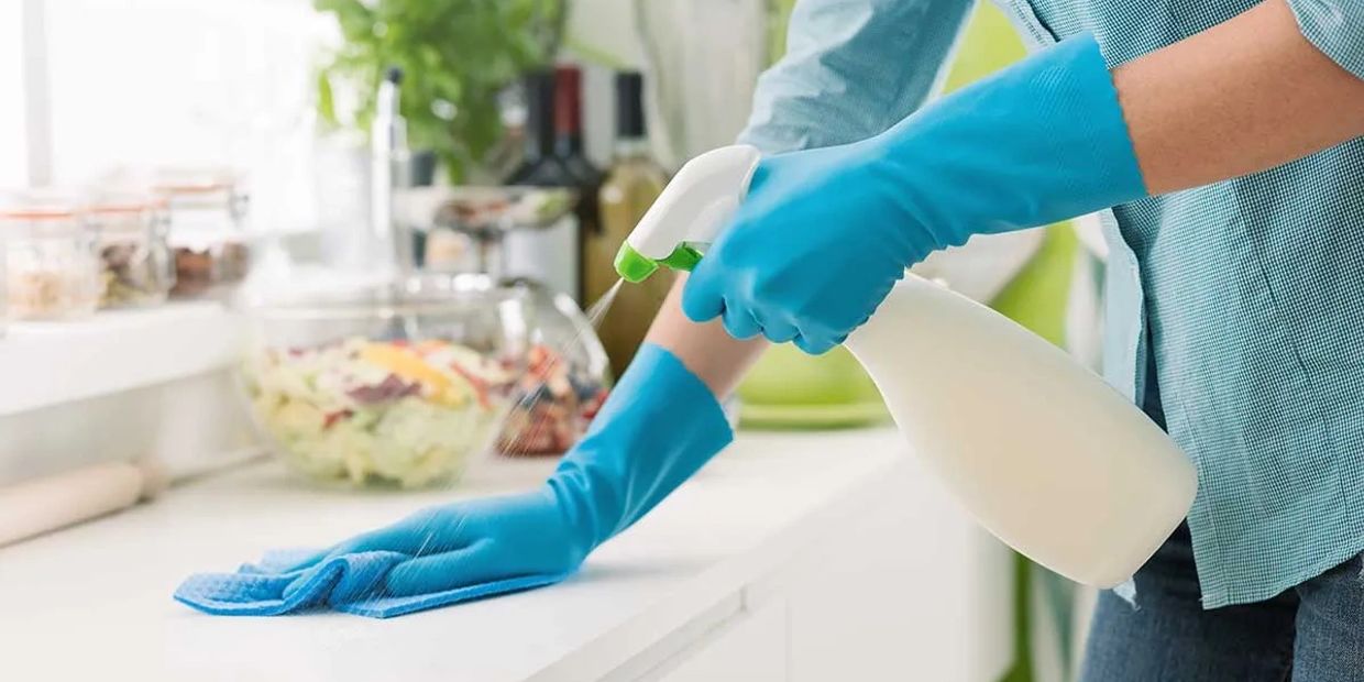 Person cleaning kitchen counter with spray bottle and cloth wearing blue gloves.