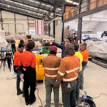 Workers in safety gear gather at an industrial workshop for a briefing or demonstration.