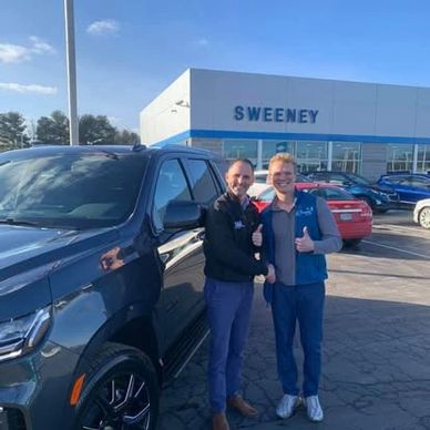 Two men smiling and giving thumbs up beside a black SUV at a car dealership.