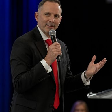 Man in suit and red tie speaking into a microphone on stage.