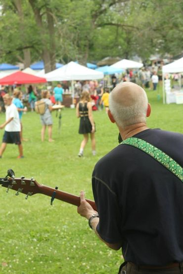 Bald man playing guitar at an outdoor festival with people and tents in the background.