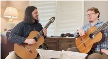 Two men playing acoustic guitars and smiling indoors.
