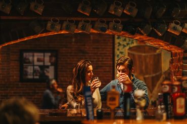 Two people sit and converse in a dimly lit bar with exposed brick walls and ambient lighting.