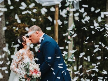 Wedding couple kissing under falling white confetti against dark background.