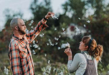A couple in casual clothing blowing dandelions together in a garden during golden hour.