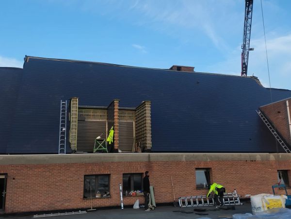 Workers on a brick building roof installing dark roofing material under a clear blue sky.