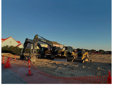 Construction equipment parked on a sandy site with houses in the background.