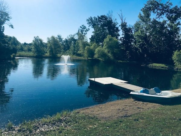 A serene pond with a fountain, a wooden dock, and a paddle boat on the grassy bank under a clear sky