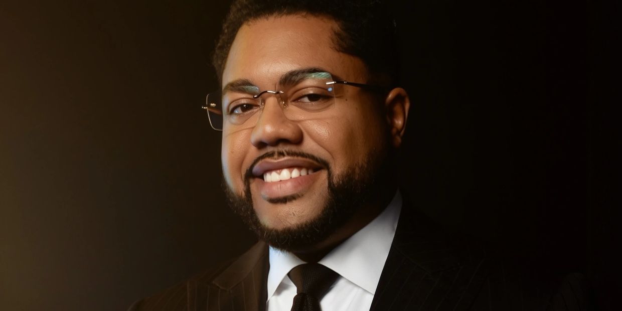 Confident man wearing glasses and a suit smiling warmly against a dark background.