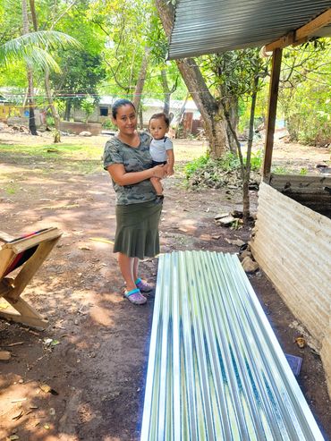 Woman holding a child outdoors near a metal sheet under a roof.