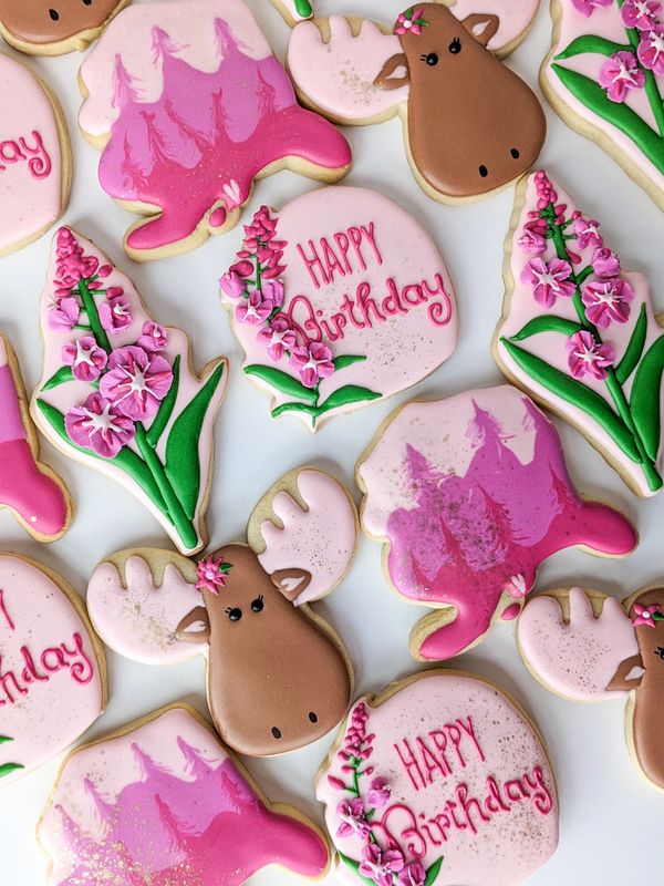 Pink-themed sugar cookies shaped like moose, Alaska, fireweed, and happy birthday plaques