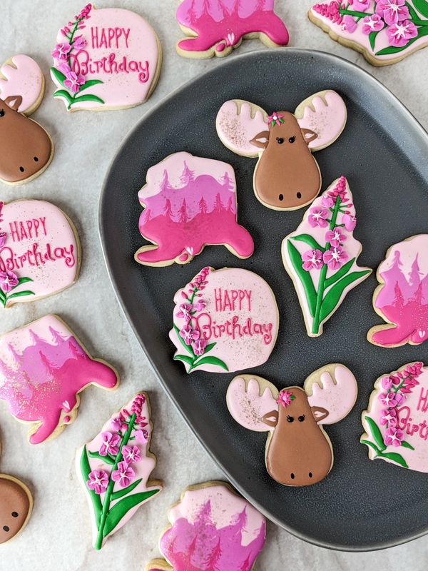 Pink-themed sugar cookies shaped like moose, Alaska, fireweed, and happy birthday plaques on a plate