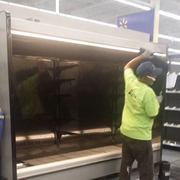 A worker wearing gloves and a mask installs or adjusts a large black refrigerated display case.
