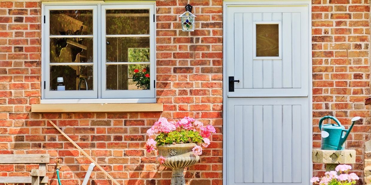 Brick house with white door, pink flowers in a planter, green watering can, white framed window