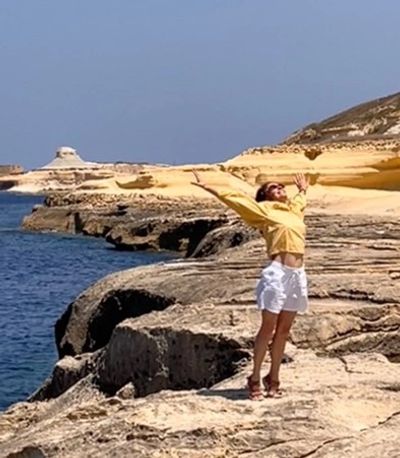 A woman wearing a yellow top and white shorts stands on a sunny, rocky coastal cliff