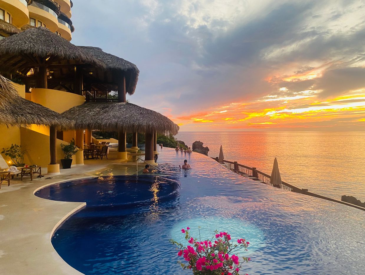 Infinity pool at sunset with tropical thatched roofs and ocean view.
