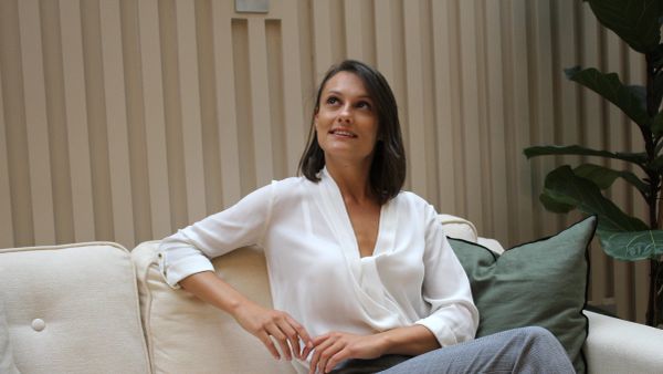 Woman in white blouse sitting on a beige sofa, looking up thoughtfully.