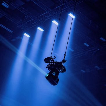 A camera suspended by cables under blue stage lights.