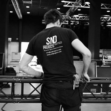 Man setting up stage equipment wearing an SND Projects t-shirt.