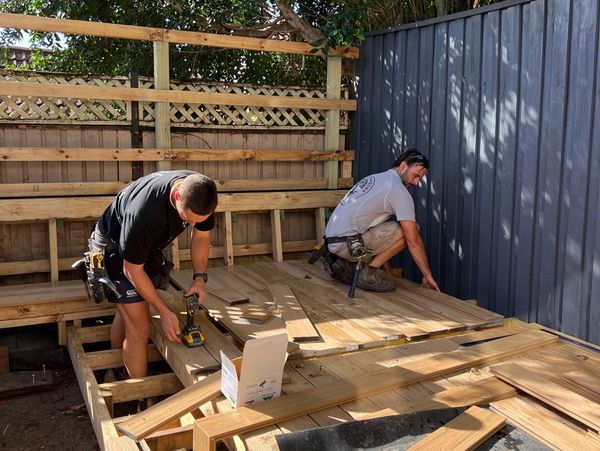 Two workers building a wooden deck in a backyard under trees.