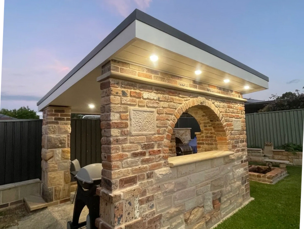 Brick outdoor kitchen with built-in grill and overhead lights at dusk.