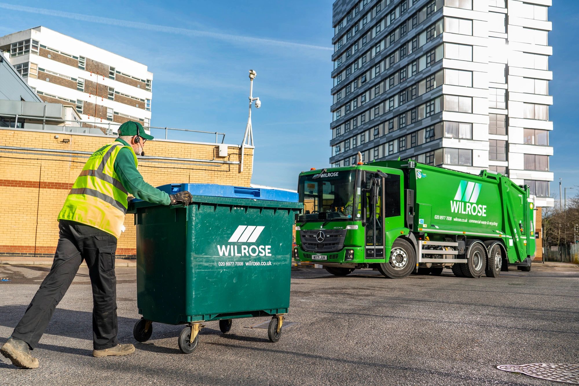 wilrose 1100L wheelie bin being moved by driver with green dustcart in the background.