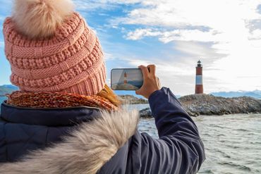 Person in winter clothing photographing a lighthouse on rocky coast with smartphone.