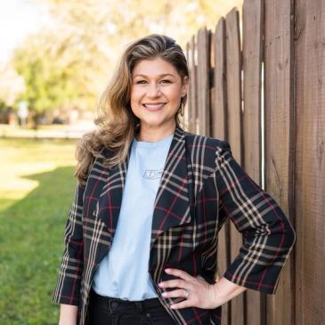 Smiling woman in plaid blazer posing by a wooden fence outdoors.