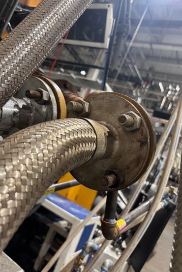 Close-up of braided metal hoses connected to a flange in an industrial setting.