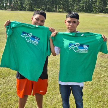Two boys holding green camp t-shirts outdoors on a sunny day.