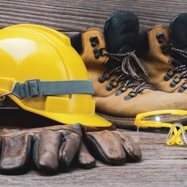 Safety gear including a yellow hard hat, gloves, boots, and protective glasses on wooden surface.