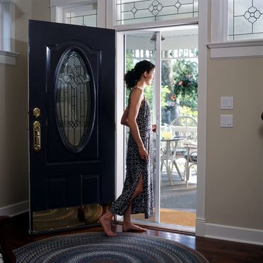 Woman standing barefoot by an open front door leading to a sunny porch.