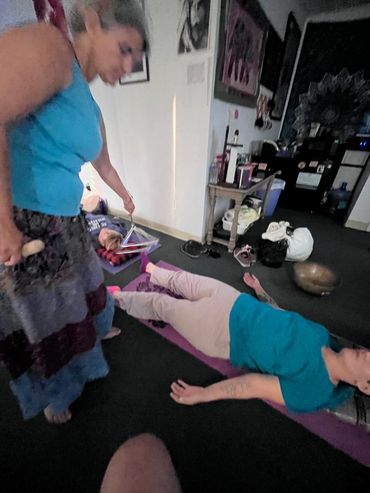 A woman lying on a mat while another woman stands nearby holding a wooden object.