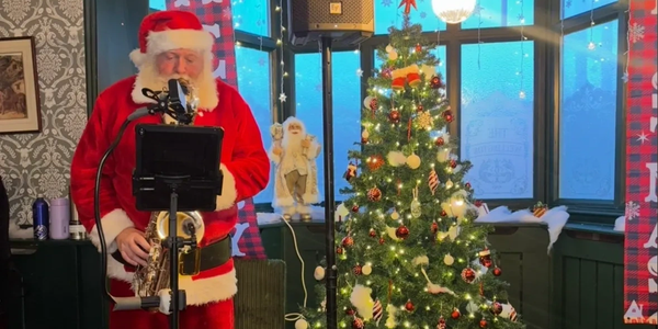 Santa Claus playing saxophone by a decorated Christmas tree indoors.