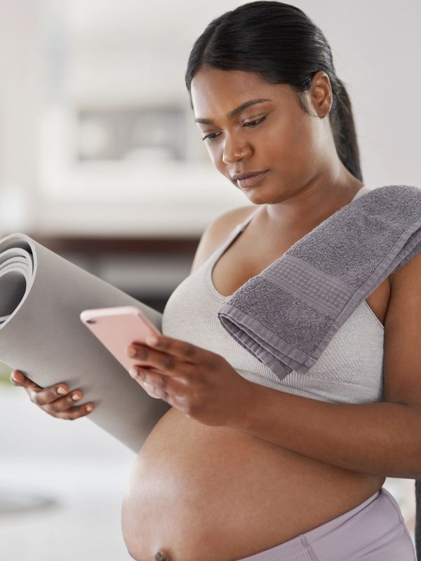 a pregnant woman looks at her phone while holding a yoga mat