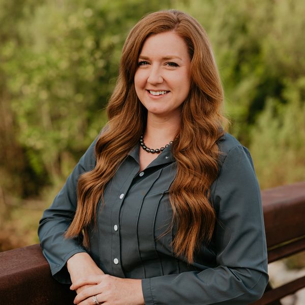 Smiling woman with long red hair posing outdoors in a gray shirt and black necklace.