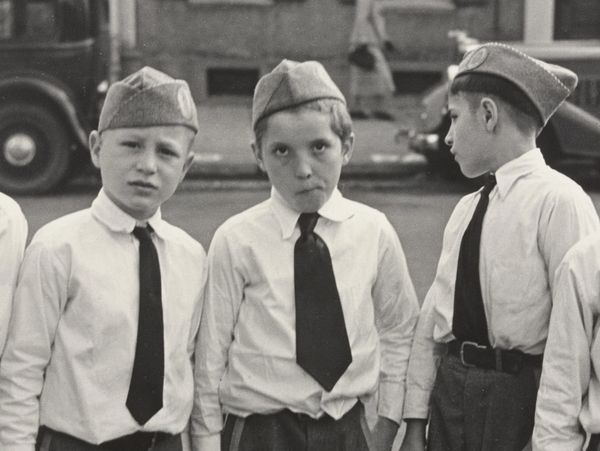 Three boys in uniform shirts, ties, and caps, standing outdoors with a vintage car in the background.