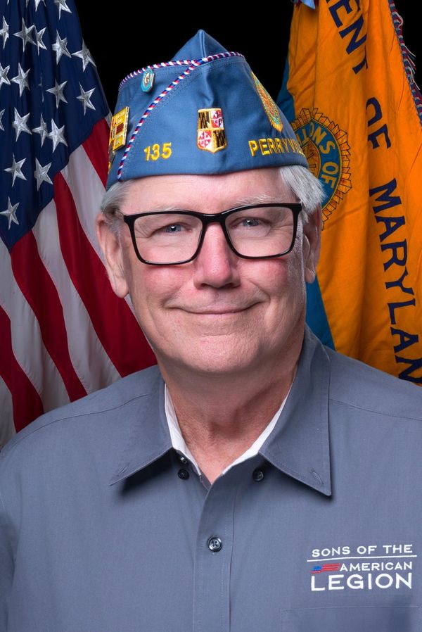 Smiling man wearing a Sons of the American Legion uniform and cap, standing in front of American and Maryland flags.