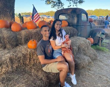 Couple with baby sitting on hay bales surrounded by pumpkins and an American flag.