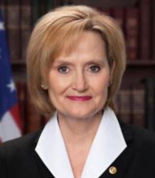 Professional portrait of a woman with short blonde hair and an American flag in the background.