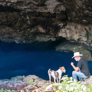 A man in a cowboy hat sits with his dog at the entrance to a cenote, overlooking deep blue water,
