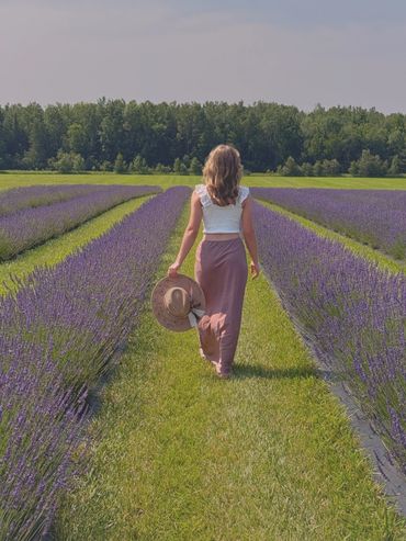 Woman walking through a lavender field holding a hat under a clear sky.