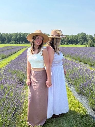 Two women holding hands in a lavender field, wearing hats and flowing skirts.
