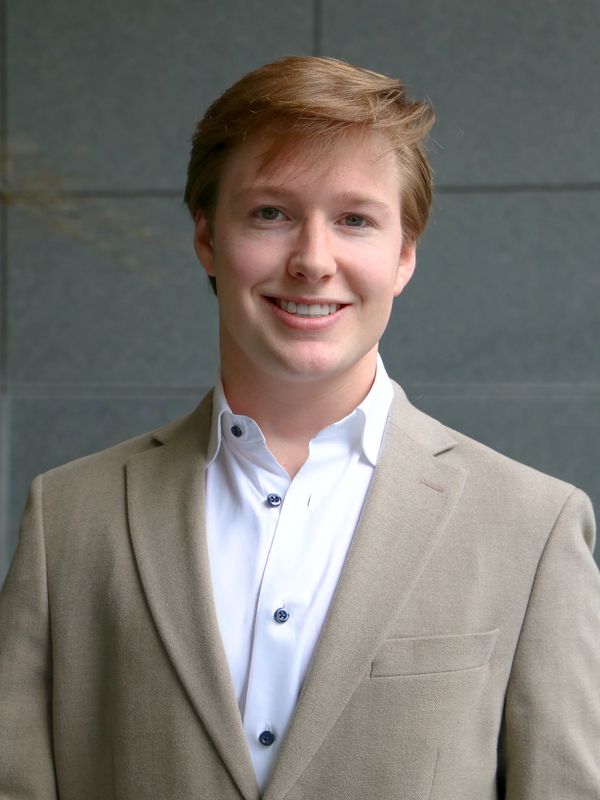 Young man in beige blazer smiling against a gray wall.