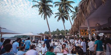 Crowded outdoor beachside restaurant with palm trees and people dining.